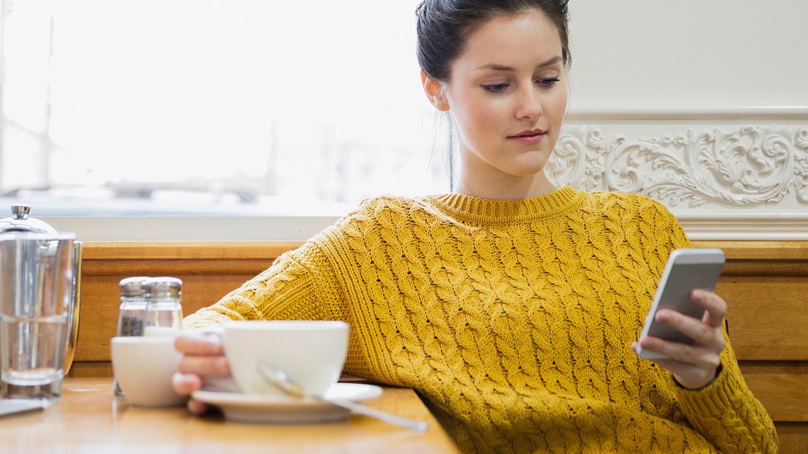 A woman sitting in a cafe holding a cup in right hand and viewing a mobile phone held in her left hand.