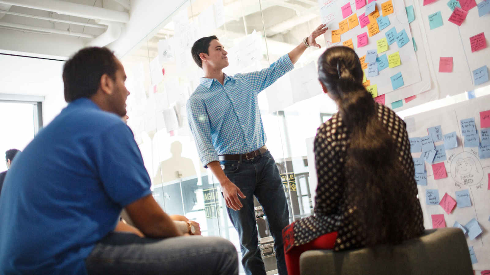 Man gesturing to a board with several post-it notes while two people look on.